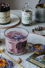 A lavender-colored beverage in a vintage glass with purple flower details, styled on a white lace runner with dried flowers, a card, and bottles in the background.