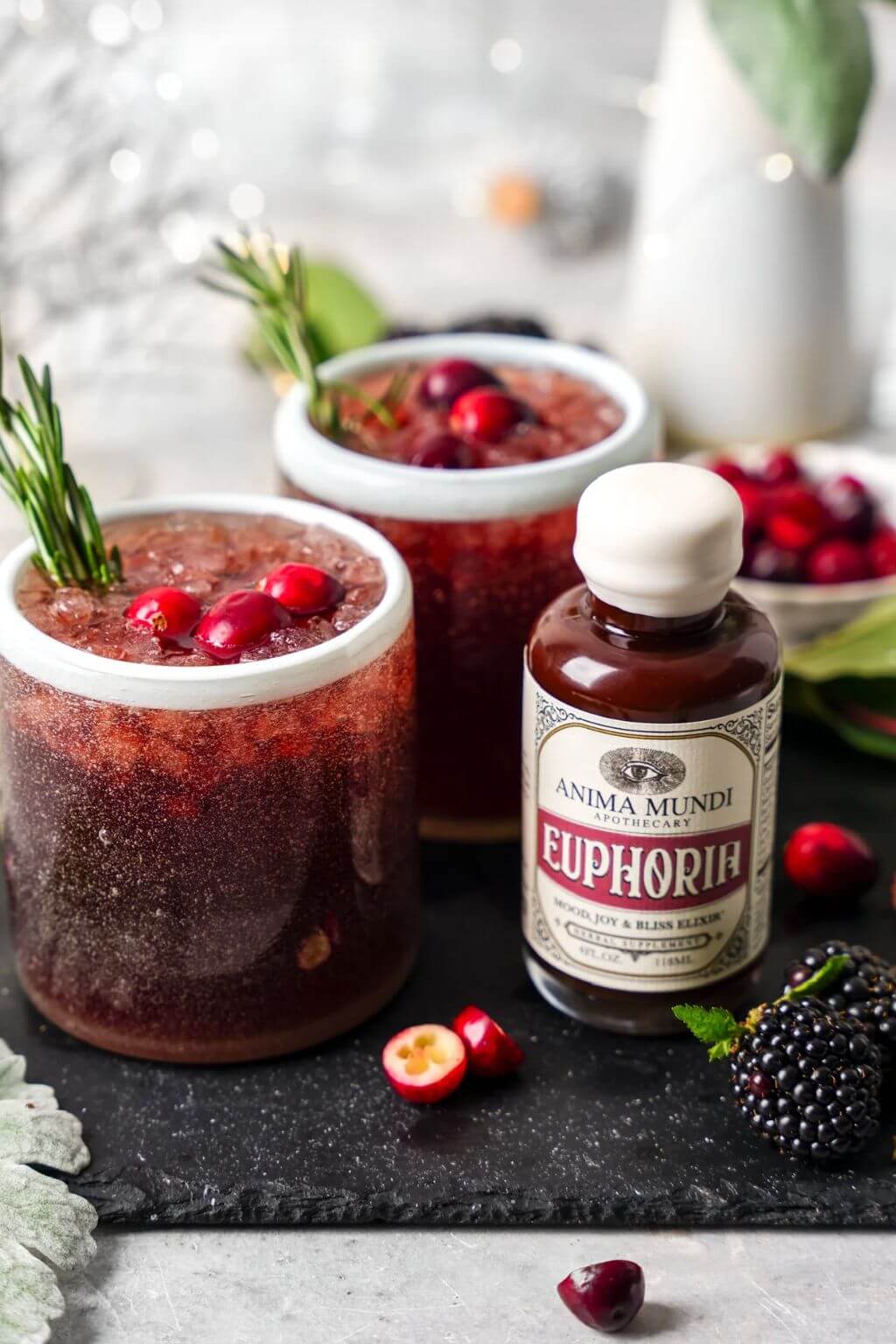 Two glasses of dark red mocktails with fresh cranberries and rosemary garnish, alongside a bottle of Anima Mundi Euphoria syrup on a dark slate board with blackberries and cranberries.