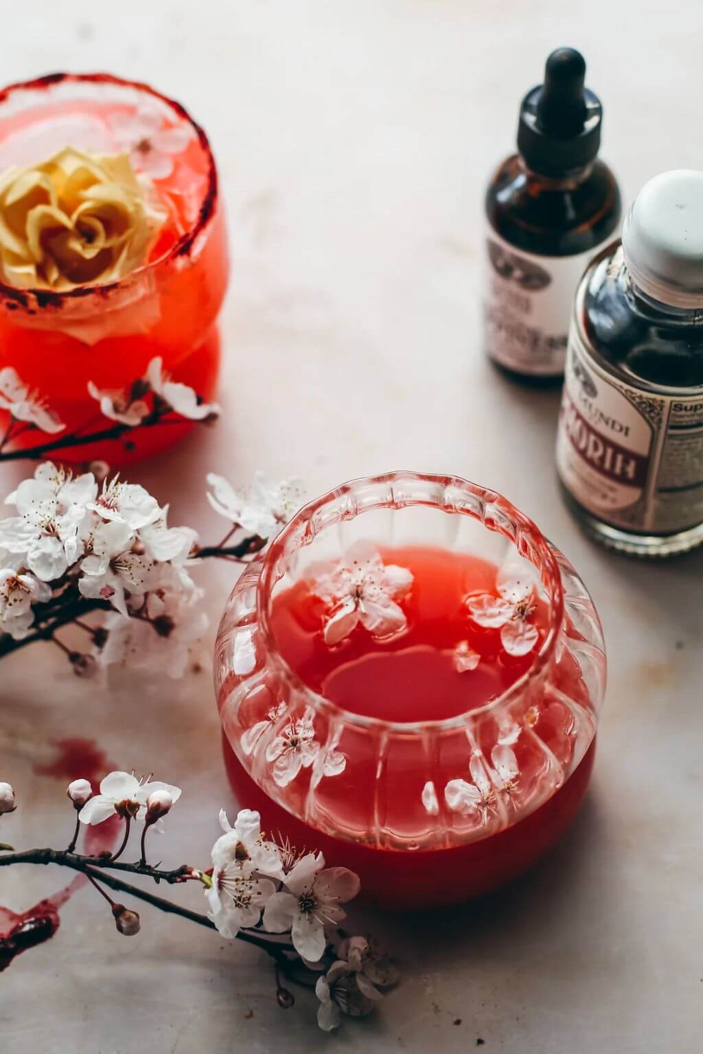 A styled tabletop scene with two glass cups containing vibrant red beverages, one with a gold flower on top and the other with white flower petal. Small bottles with dropper and cap closures sit behind, and white blossoms are arranged around the glasses on a light surface.