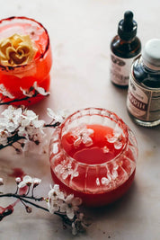 A styled tabletop scene with two glass cups containing vibrant red beverages, one with a gold flower on top and the other with white flower petal. Small bottles with dropper and cap closures sit behind, and white blossoms are arranged around the glasses on a light surface.