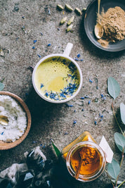 A styled flat-lay of a cup of golden moon milk garnished with blue lotus flowers, surrounded by ingredients including a bowl of white salt, a jar of turmeric powder, green cardamom pods, and eucalyptus leaves on a textured stone surface.