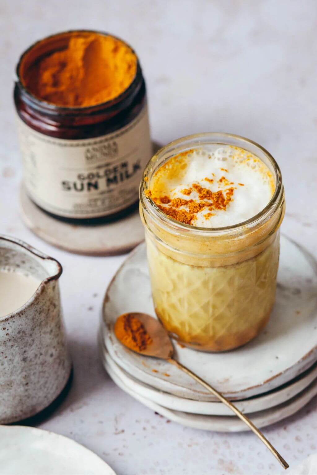 A styled product shot of a glass jar labeled 'Golden Sun Milk' filled with a yellow-orange beverage topped with white foam and a sprinkle of turmeric powder, alongside a small cream-colored pitcher and spoon on stacked plates.