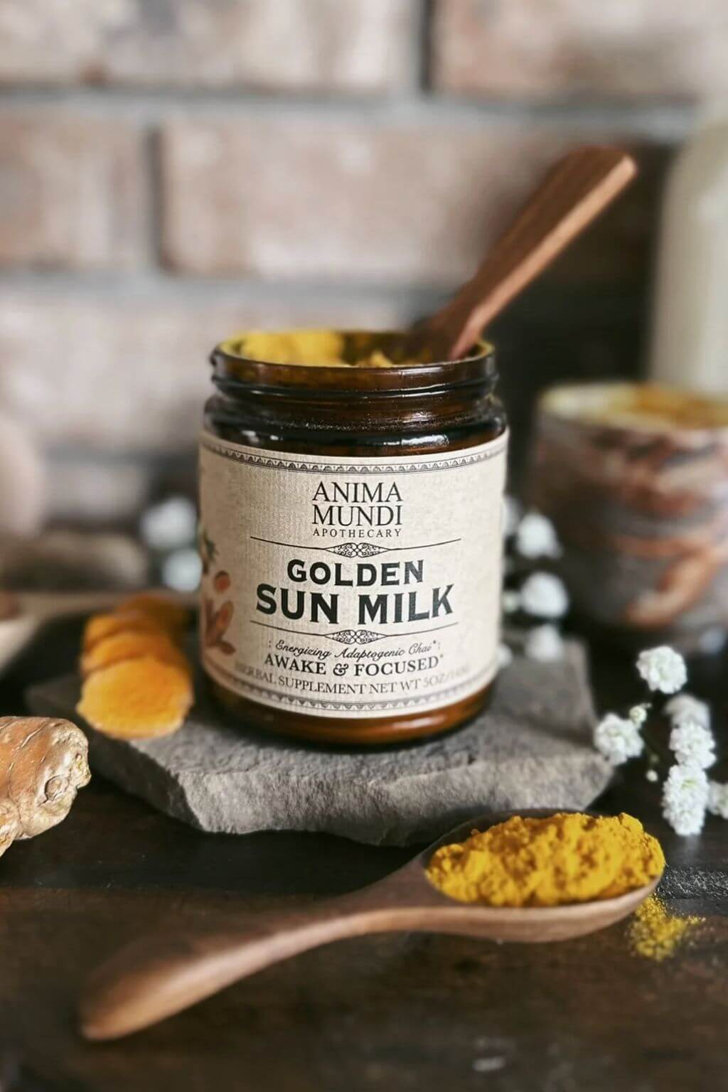 A jar of Anima Mundi Apothecary Golden Sun Milk with a wooden spoon inside, set on a dark surface with a stone slab, ginger root, white flowers, and a wooden spoon of turmeric powder in front, against a blurred brick wall background.