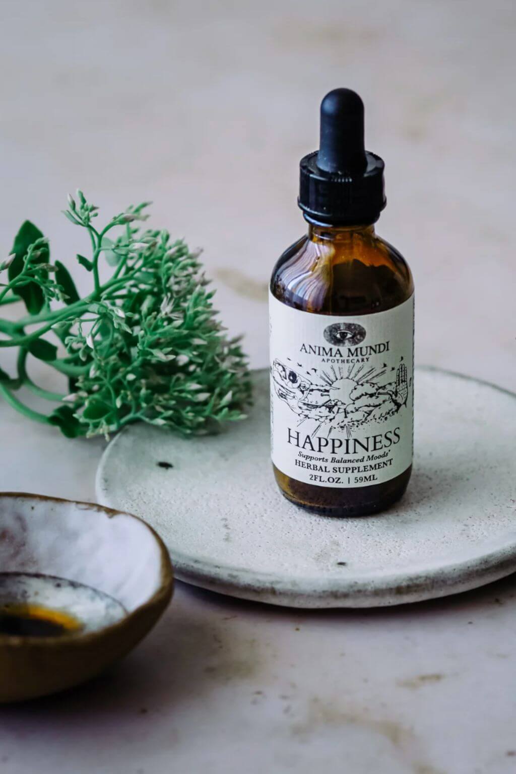 Bottle of Anima Mundi's Happiness Tincture on a ceramic plate with greenery and muted gray background