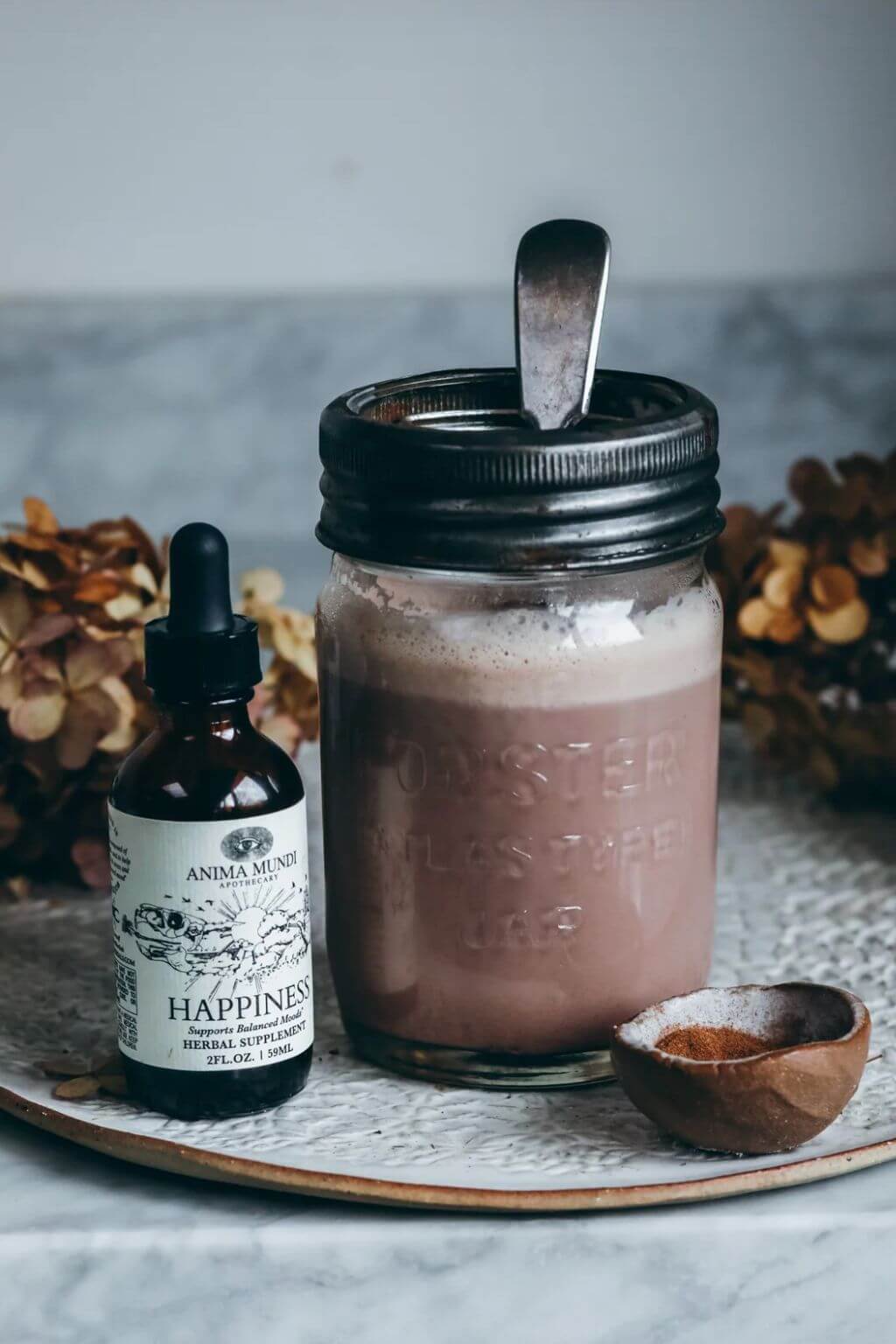 A mason jar filled with a frothy chocolate beverage, placed beside a small amber dropper bottle labeled Anima Mundi Happiness Tonic on a tray with dried flowers and a small bowl of cinnamon.