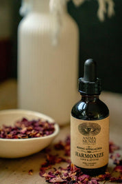 A small amber glass dropper bottle labeled Anima Mundi Harmonize Love & Affection, styled on a tabletop with dried rose petals in a bowl and a blurred white bottle in the background.
