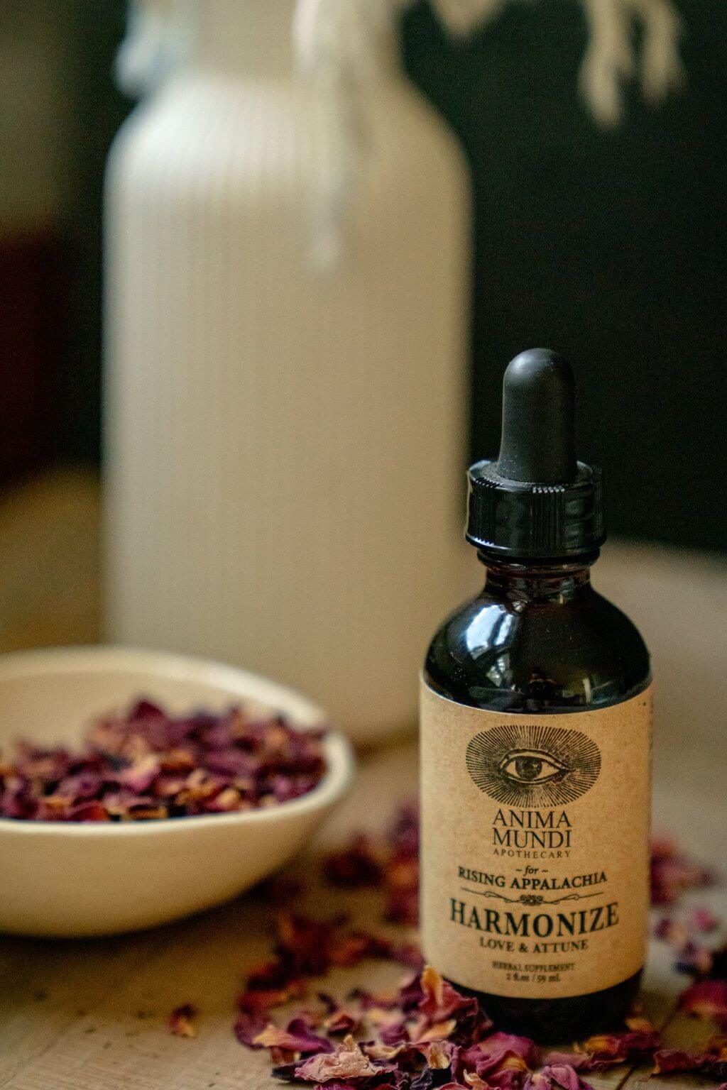 A small amber glass dropper bottle labeled Anima Mundi Harmonize Love & Affection, styled on a tabletop with dried rose petals in a bowl and a blurred white bottle in the background.