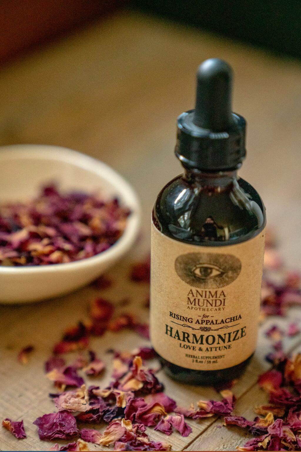 A small amber glass dropper bottle labeled Anima Mundi Harmonize Tonic on a wooden surface with scattered dried rose petals and a bowl of dried petals in the background.