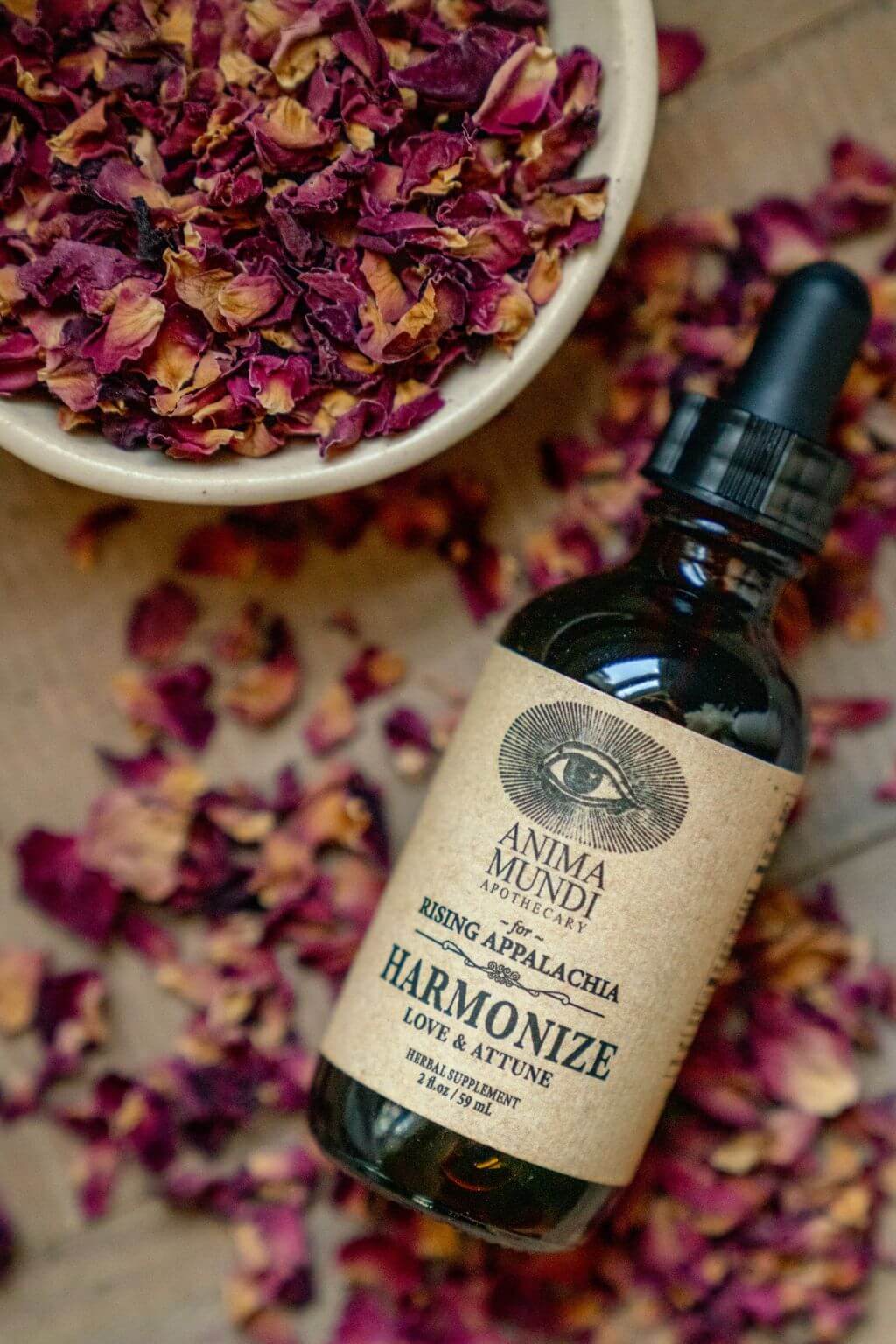 A dark glass dropper bottle labeled Anima Mundi Rising Appalachia Harmonize Love & Attune, styled on a rustic surface with dried rose petals and a bowl of loose dried roses.