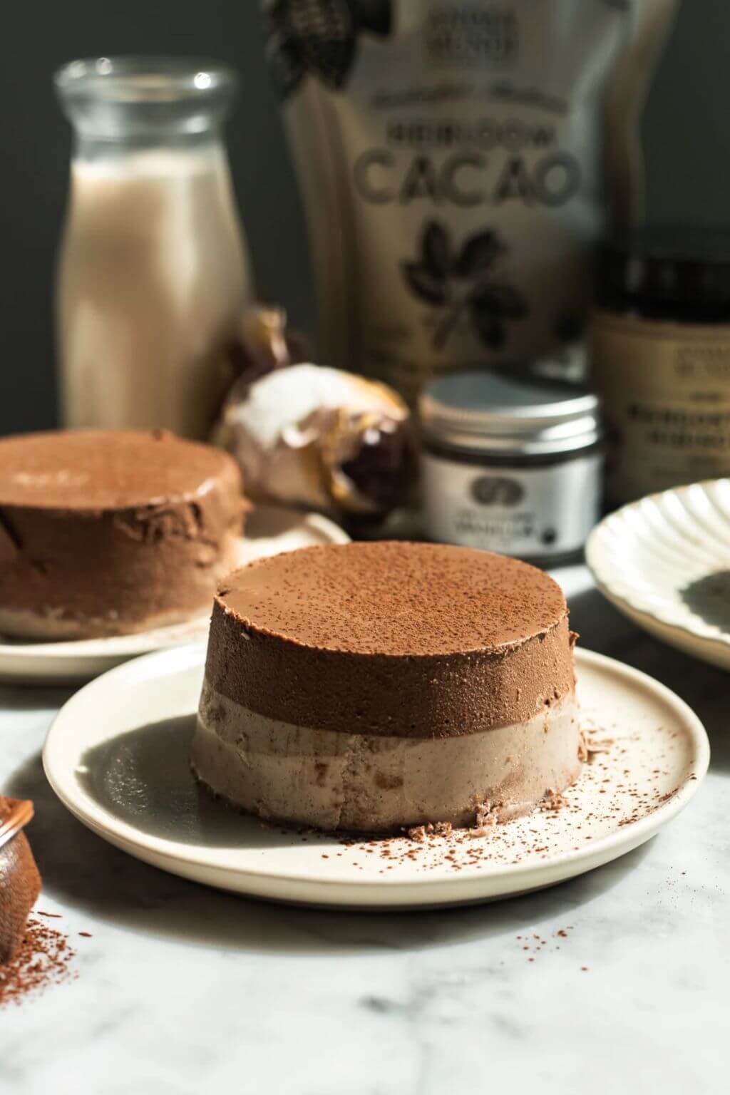 A chocolate mousse dessert on a small plate, with another dessert and ingredients like cacao powder and a milk bottle blurred in the background.