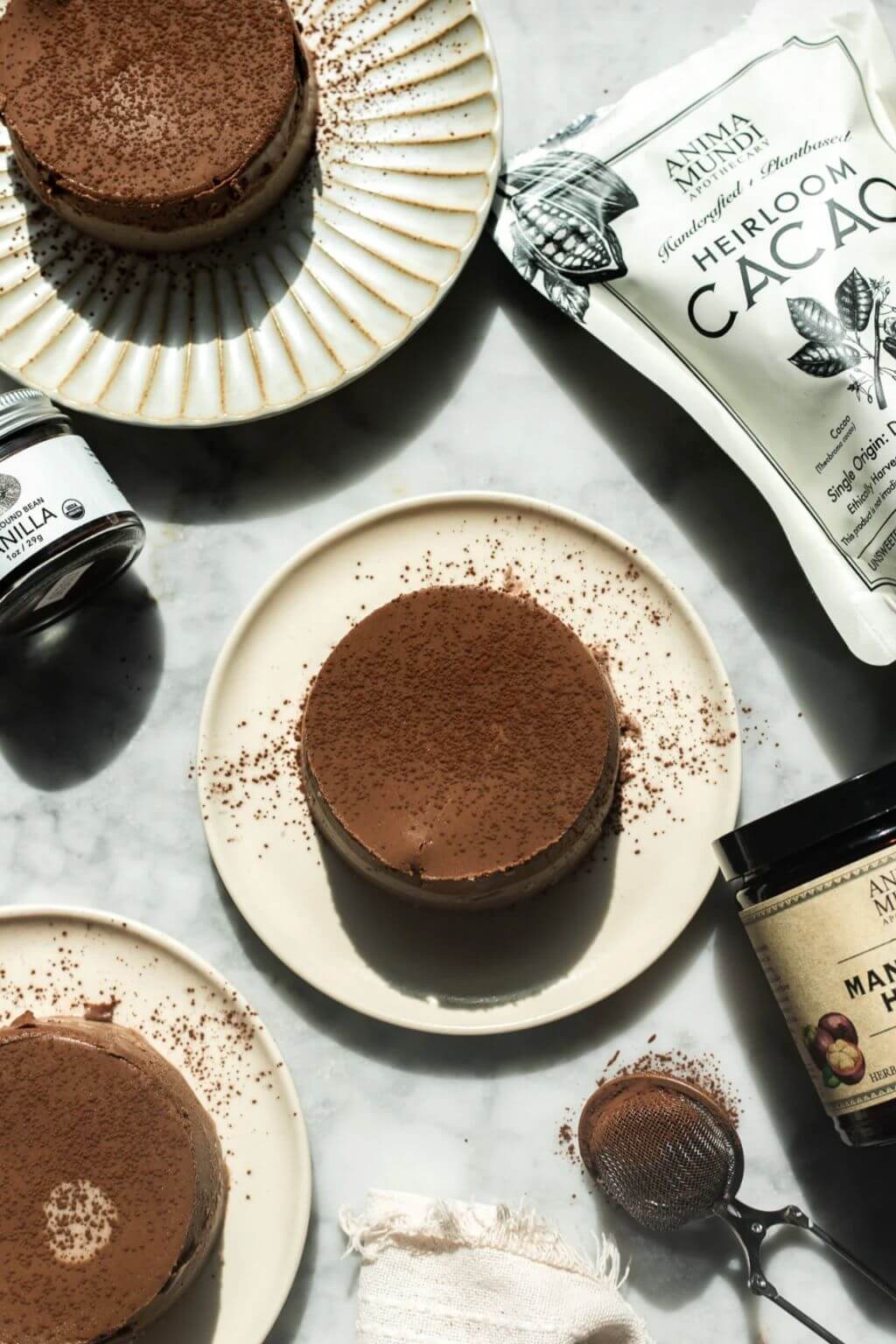 Three chocolate desserts on cream-colored plates, arranged on a light marble surface with cocoa powder, a pouch of Anima Mundi Heirloom Cacao, vanilla extract, and a jar of Madagascar vanilla beans.