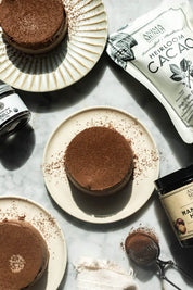 Three chocolate desserts on cream-colored plates, arranged on a light marble surface with cocoa powder, a pouch of Anima Mundi Heirloom Cacao, vanilla extract, and a jar of Madagascar vanilla beans.