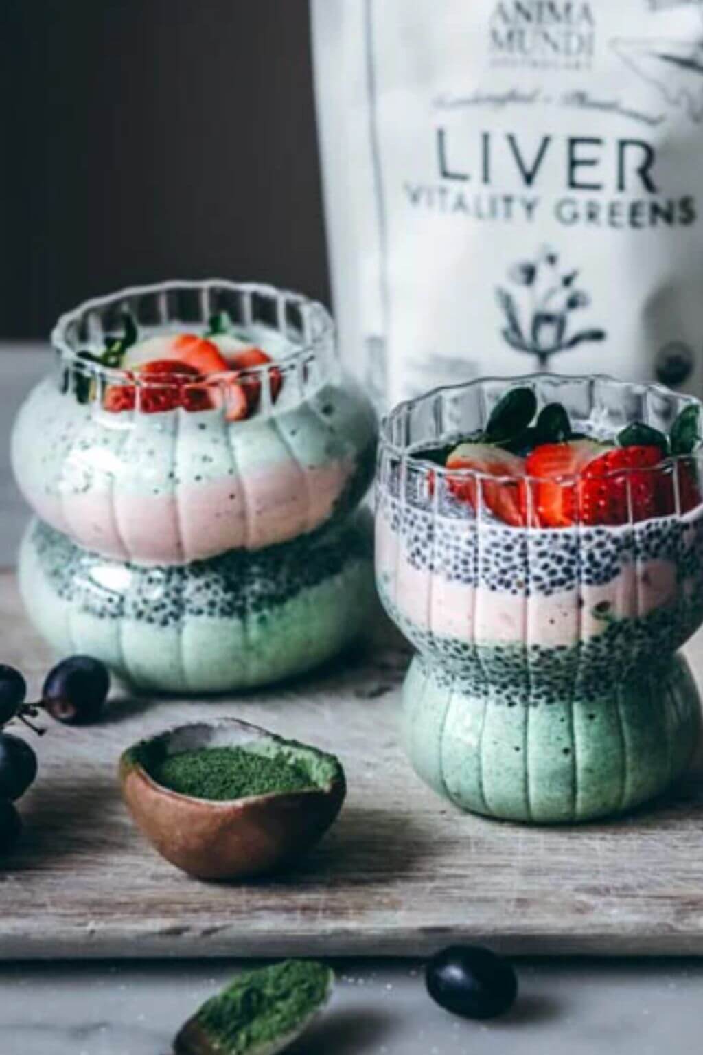 Two layered green and pink chia seed smoothies in ribbed glass cups, topped with strawberries, with a small bowl of green powder and a blurred bag labeled 'Liver Vitality Greens' in the background.
