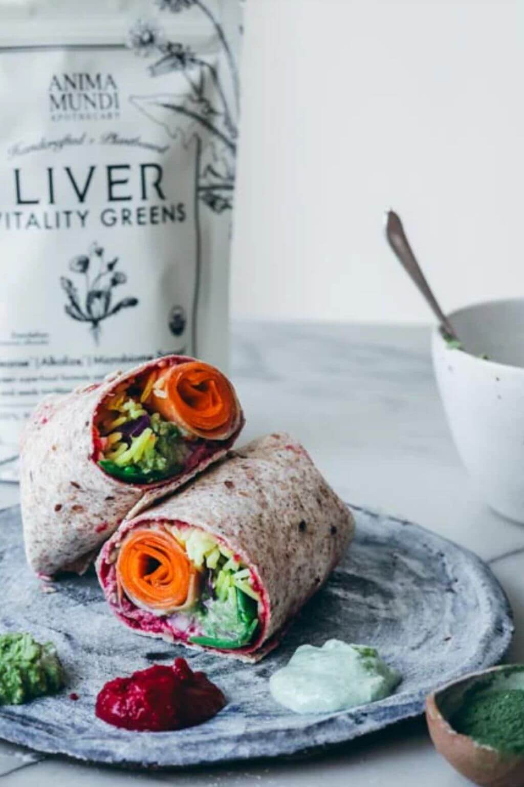 A plated wrap filled with colorful vegetables, with a bag of Anima Mundi Liver Vitality Greens in the background and a bowl of green sauce on the side.