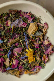 A close-up of loose-leaf herbal tea mixed with dried rose petals, yellow flower pieces, and green leaves inside a white ceramic bowl.