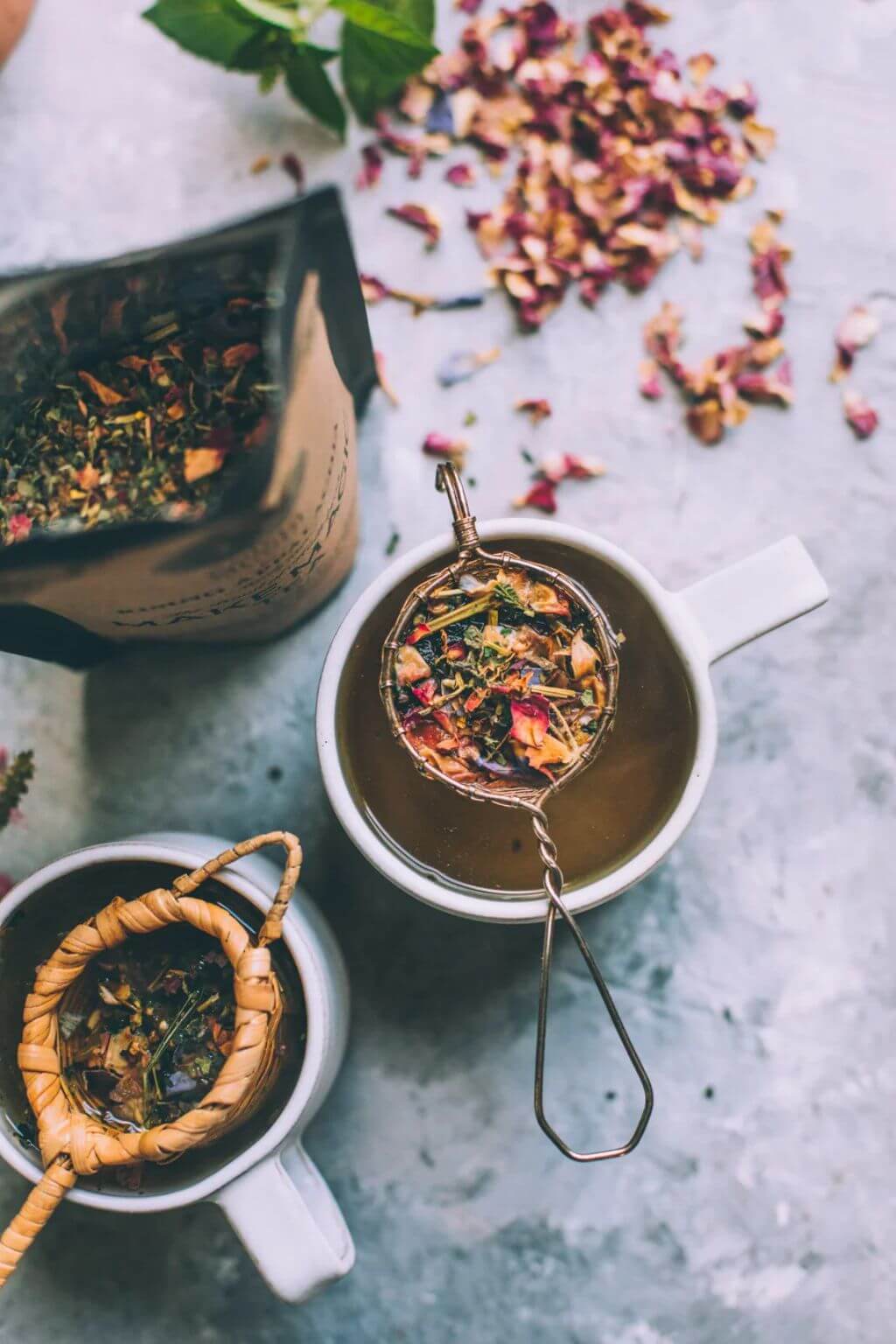 Tea-making process with loose tea leaves and both a woven brass and a bamboo tea strainer resting above white mugs atop a concrete surface with dried rose petals.