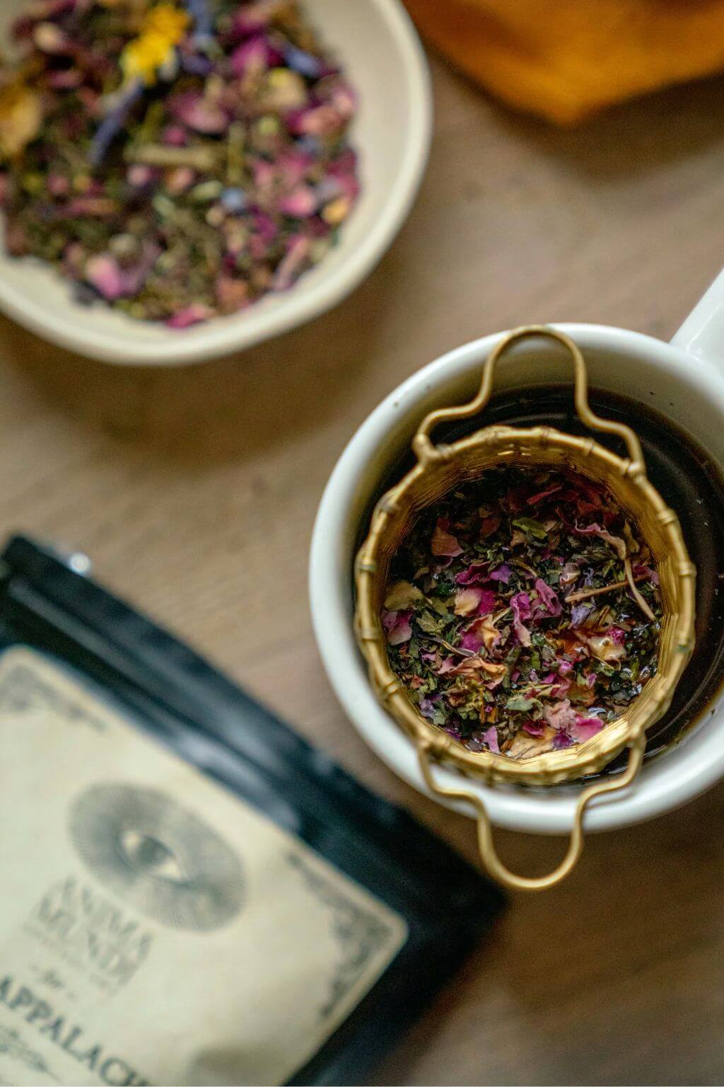A top-down view of a white ceramic mug with a woven brass infuser filled with dried floral tea leaves, alongside a bowl of loose herbal tea and a black tea pouch on a wooden surface.