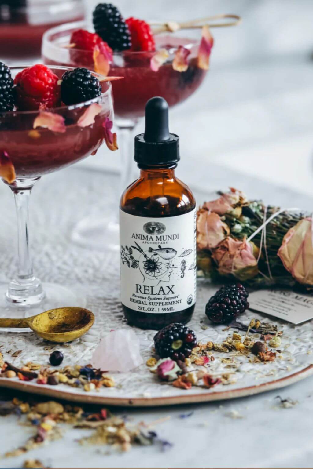 An amber glass dropper bottle labeled 'Anima Mundi Relax Herbal Supplement' sits on a tray with dried flowers, blackberries, and loose botanicals, with two pink berry cocktails blurred in the background.
