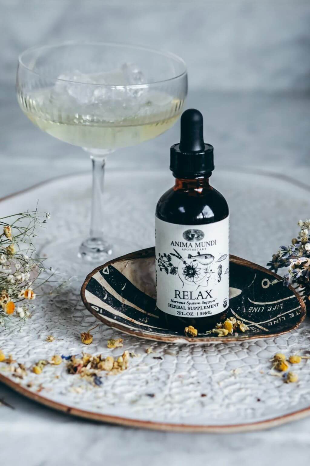 A small amber dropper bottle labeled 'ANIMA MUNDI RELAX' sits on a patterned dish with dried flowers, next to a coupe glass filled with a pale yellow drink on a textured white tray.