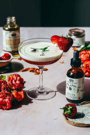 A styled tabletop scene featuring a stemmed cocktail glass filled with a layered pink drink topped with white foam and a strawberry garnish, surrounded by flowers, a bottle labeled RELAX on a pale surface.
