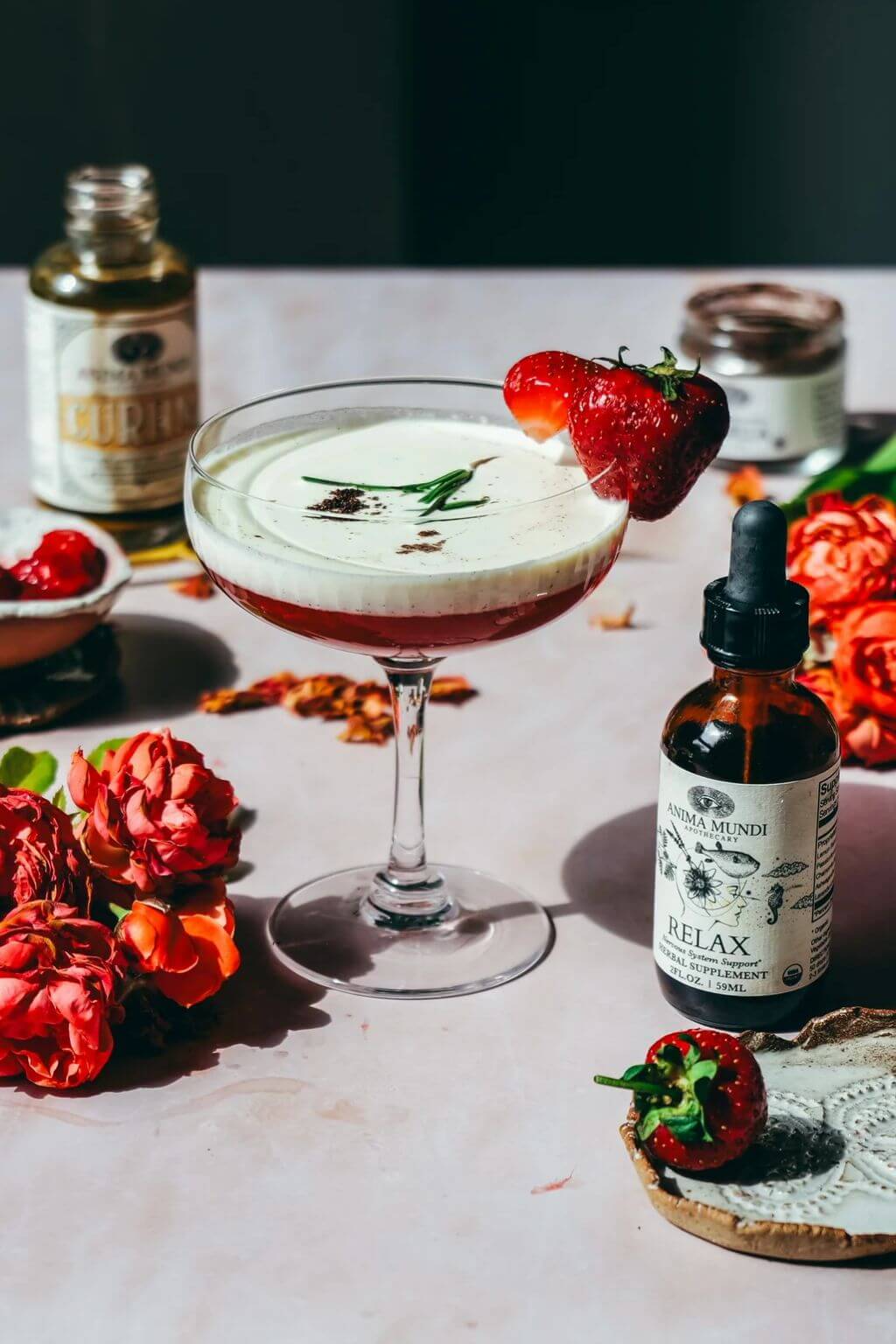 A styled tabletop scene featuring a stemmed cocktail glass filled with a layered pink drink topped with white foam and a strawberry garnish, surrounded by flowers, a bottle labeled RELAX on a pale surface.