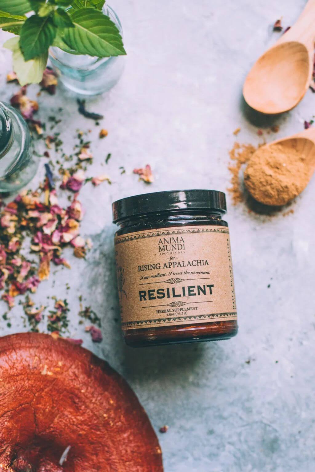 A amber glass jar of Resilient Powder by Anima Mundi + Rising Appalachia on a gray background with rose petals and wooden spoons.