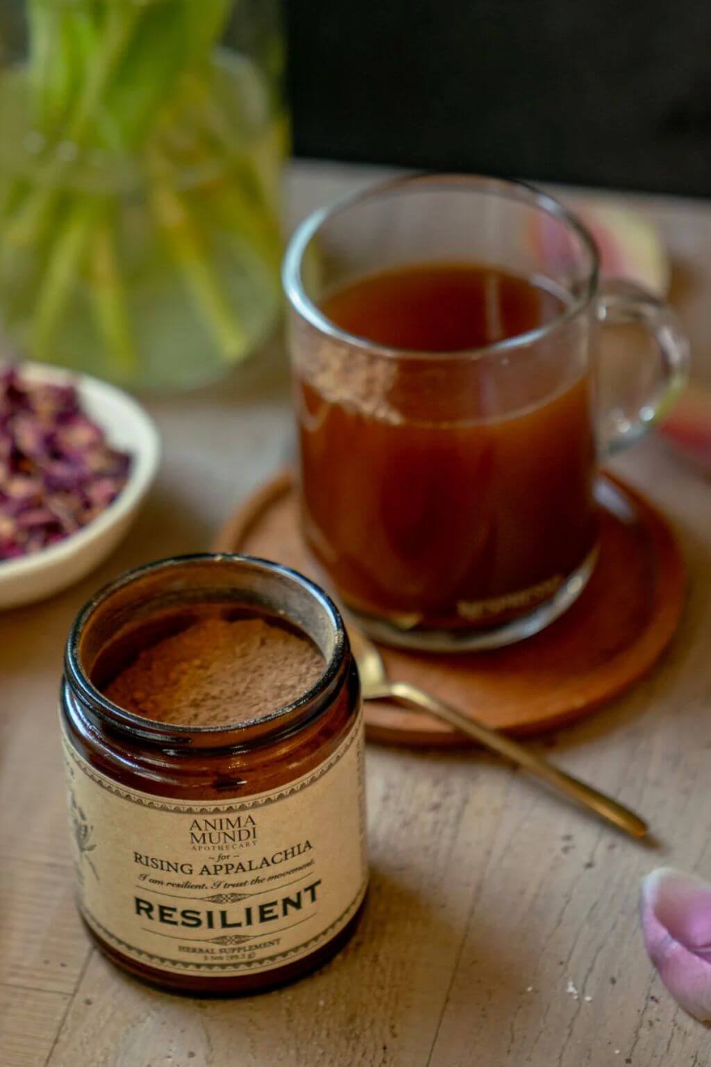 A small amber glass jar labeled 'Anima Mundi Resilient' filled with brown powder, placed on a wooden table beside a glass mug of Resilient Hot Beverage and a spoon.