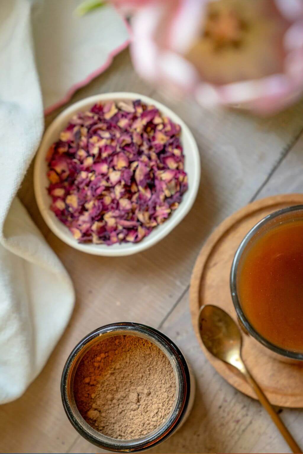 A styled flat-lay of featuring a small white bowl filled with dried pink rose petals, an open glass jar containing a Resilient Powder, a glass cup of amber liquid on a wooden saucer with a brass spoon, and a folded cream cloth on a light wood surface.