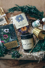 A curated gift box arrangement on a rustic wooden surface, featuring a tarot card deck box labeled 'The Herbal Astrology Card Deck' by Adriana Ayales, a jar of 'Blue Lotus' tea by Anima Mundi, a bottle of 'Dream' Elixir, and palo santo wrapped in twine, surrounded by green pine branches and dried flowers.