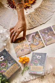 A hand reaches toward illustrated oracle cards spread on a cloth surface, with a boxed decks labeled Herbal Astrology visible in the foreground and more cards scattered in the background.