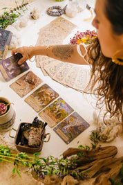 A woman with long dark hair reaches for oracle cards spread out on a white cloth, surrounded by crystals, dried flowers, herbs, and natural decor.