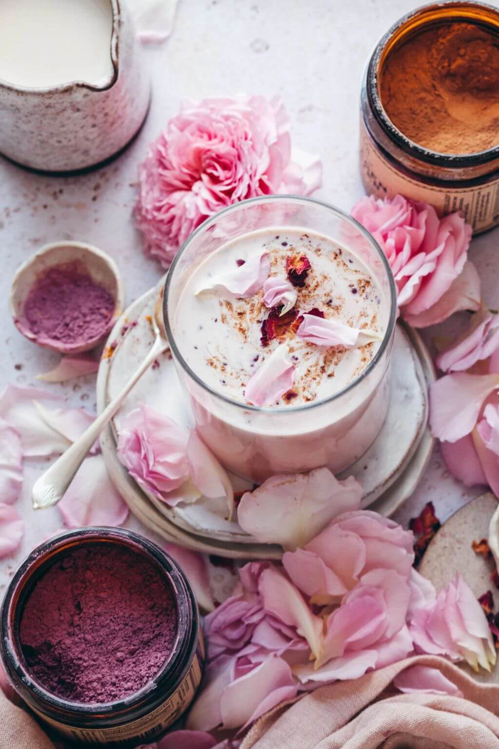 A styled flat-lay of a glass jar filled with a creamy pink beverage, surrounded by pink rose petals, loose flowers, and other open jars of matching pink and purple powders on a pale textured surface.