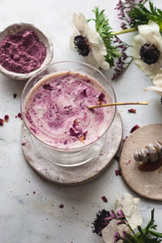 A clear glass cup filled with a creamy pink beverage, set on a round coaster with a brass spoon. The scene is styled with white and purple flowers and a small bowl of Rose Powder