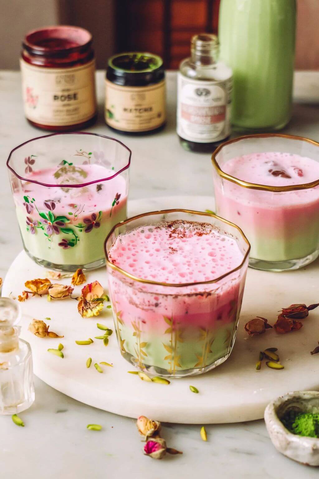 Three layered pink and green drinks in decorative glassware arranged on a white marble tray, with apothecary bottles in the blurred background.