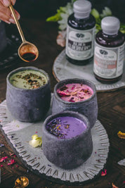 Three dark gray ceramic teacups filled with colorful herbal beverages in green, pink, and purple, arranged on a decorative tray with two bottles and a brass spoon in the background.
