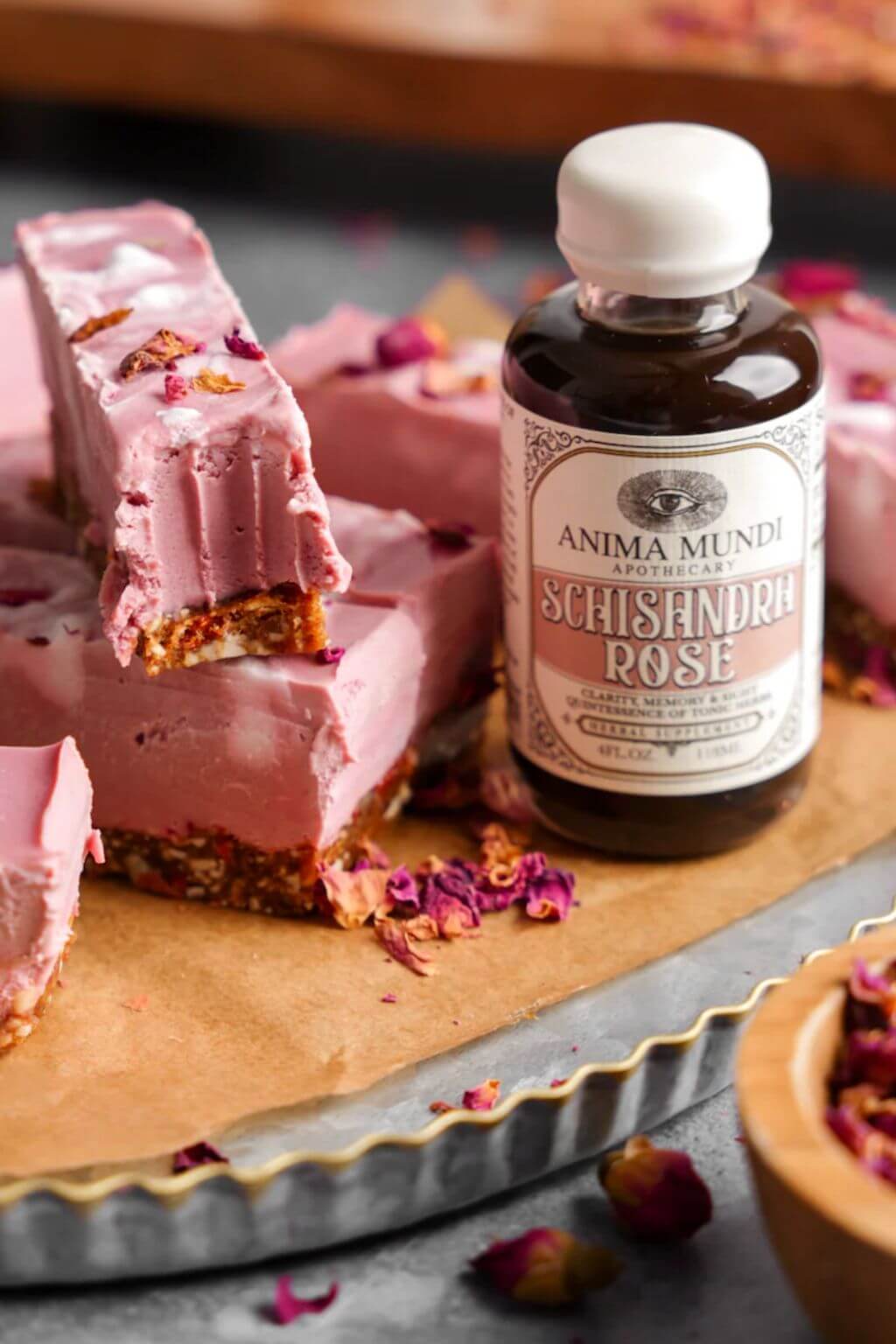 Pink frozen dessert bars with a rose-flavored base, arranged on parchment paper beside a bottle of Anima Mundi Schisandria Rose Elxiir, with scattered dried rose petals.