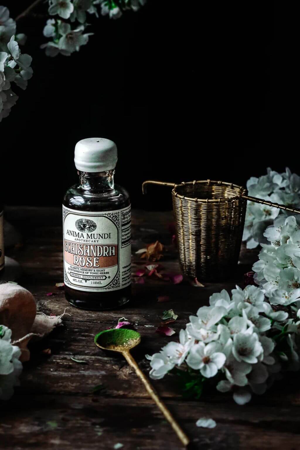 A bottle of Anima Mundi Apothecary's Schisandra Rose Elixir, styled on a rustic wooden surface with white flowers, a brass spoon, and a woven brass tea strainer against a dark background.