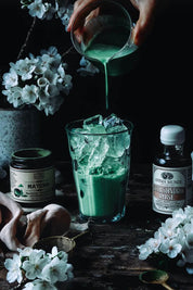 A hand pours a vibrant green matcha from a pitcher into a glass of ice on a dark rustic table. Surrounding the drink are a jar of matcha powder, a bottle of Schisandra Rose Elixir, and white cherry blossoms.