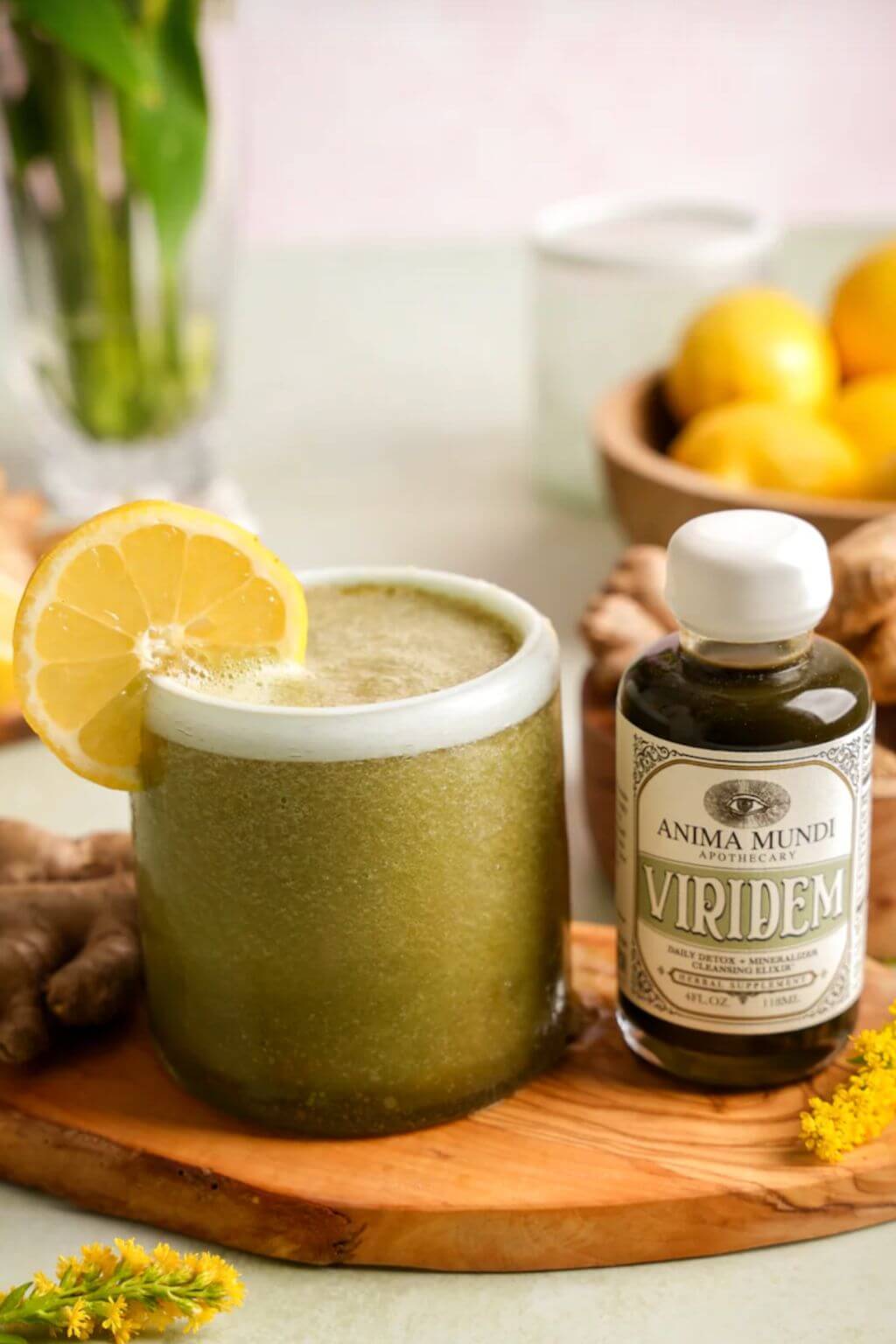 A frothy green beverage in a white-rimmed glass with a lemon slice garnish, placed on a wooden board beside a bottle of Anima Mundi Viridem Elixir, with ginger roots and lemons in the background.