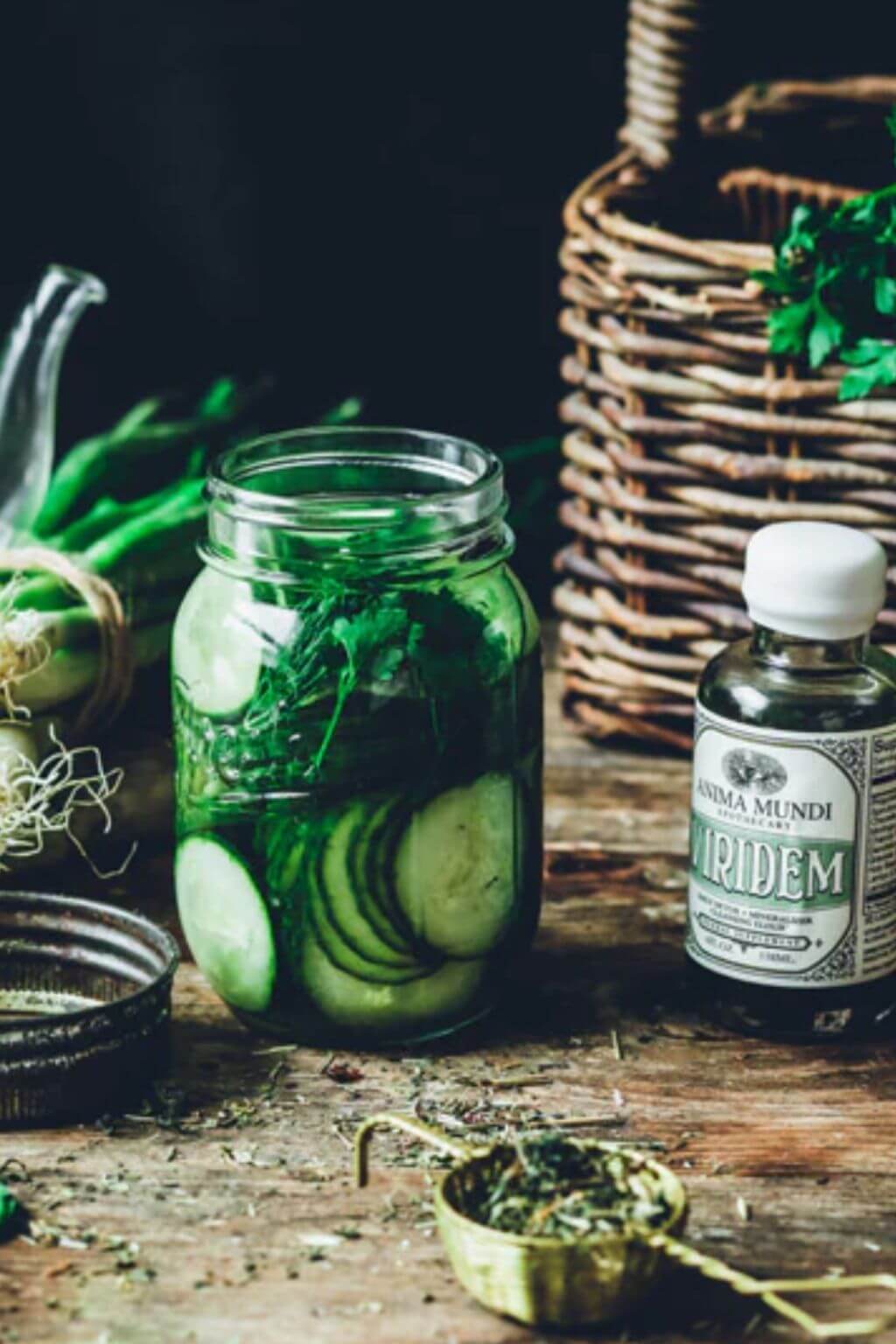 A glass mason jar filled with sliced cucumbers and green herbs sits on a rustic wooden table beside a bottle labeled 'Anima Mundi' and 'Viridem', with a wicker basket and green onions in the background.