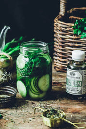 A glass mason jar filled with sliced cucumbers and green herbs sits on a rustic wooden table beside a bottle labeled 'Anima Mundi' and 'Viridem', with a wicker basket and green onions in the background.