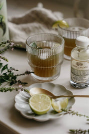 A styled tabletop scene with two ribbed clear glasses containing a green drink, a small bottle labeled 'Anima Mundi Viridem', a scalloped plate with lime wedges and a spoon, and soft neutral background elements.