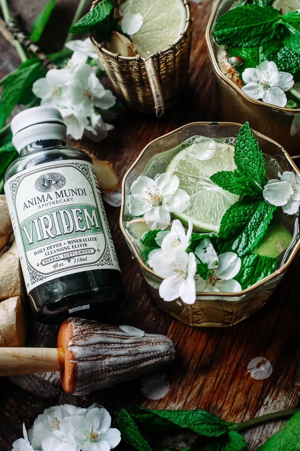 A bottle of Anima Mundi Apothecary Viridem herbal tincture is displayed next to two gol-rimmed cocktail glasses filled with a mint and lime mojito, decorated with white blossoms, on a rustic wooden table.