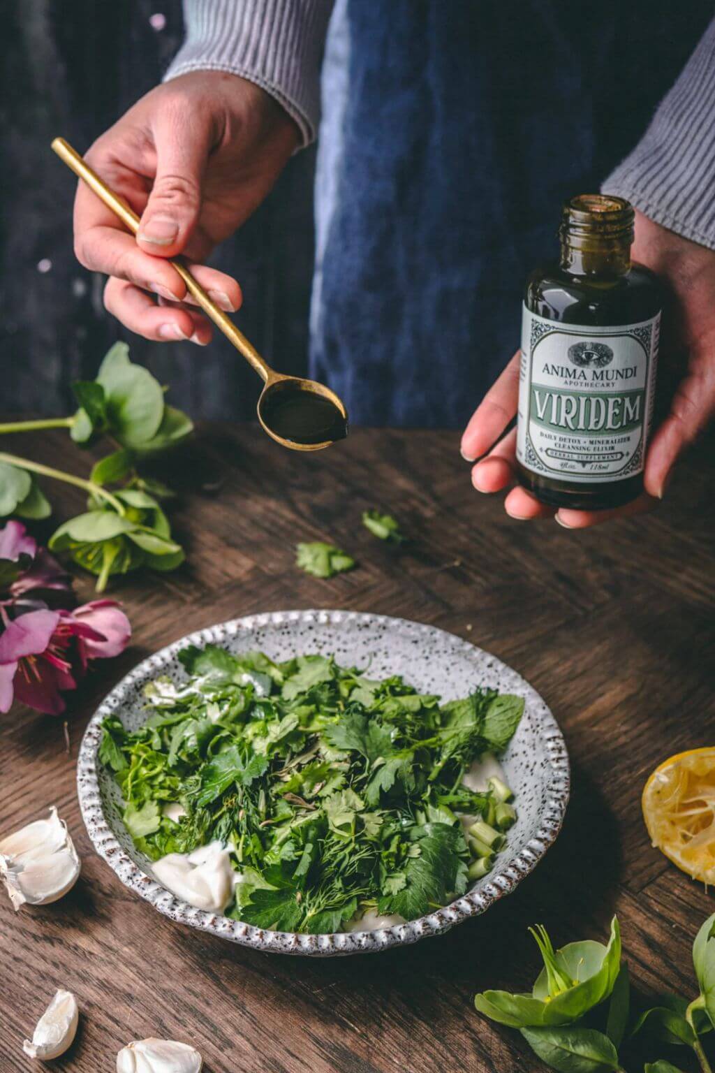 A person holds a small bottle labeled 'Anima Mundi Viridem' and a brass spoon over a bowl of chopped green herbs on a rustic wooden table.