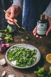 A person holds a small bottle labeled 'Anima Mundi Viridem' and a brass spoon over a bowl of chopped green herbs on a rustic wooden table.