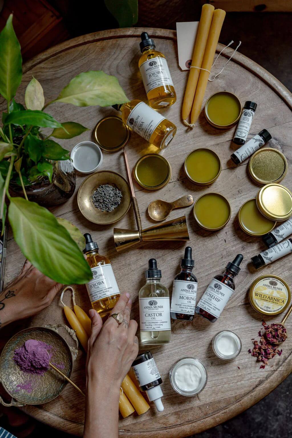 An overhead styled flat-lay of a round wooden table filled with artisanal skincare products, including dropper bottles, tins of balm, beeswax candles, small bowls, and a hand reaching in. The scene is decorated with green leaves and warm natural tones.