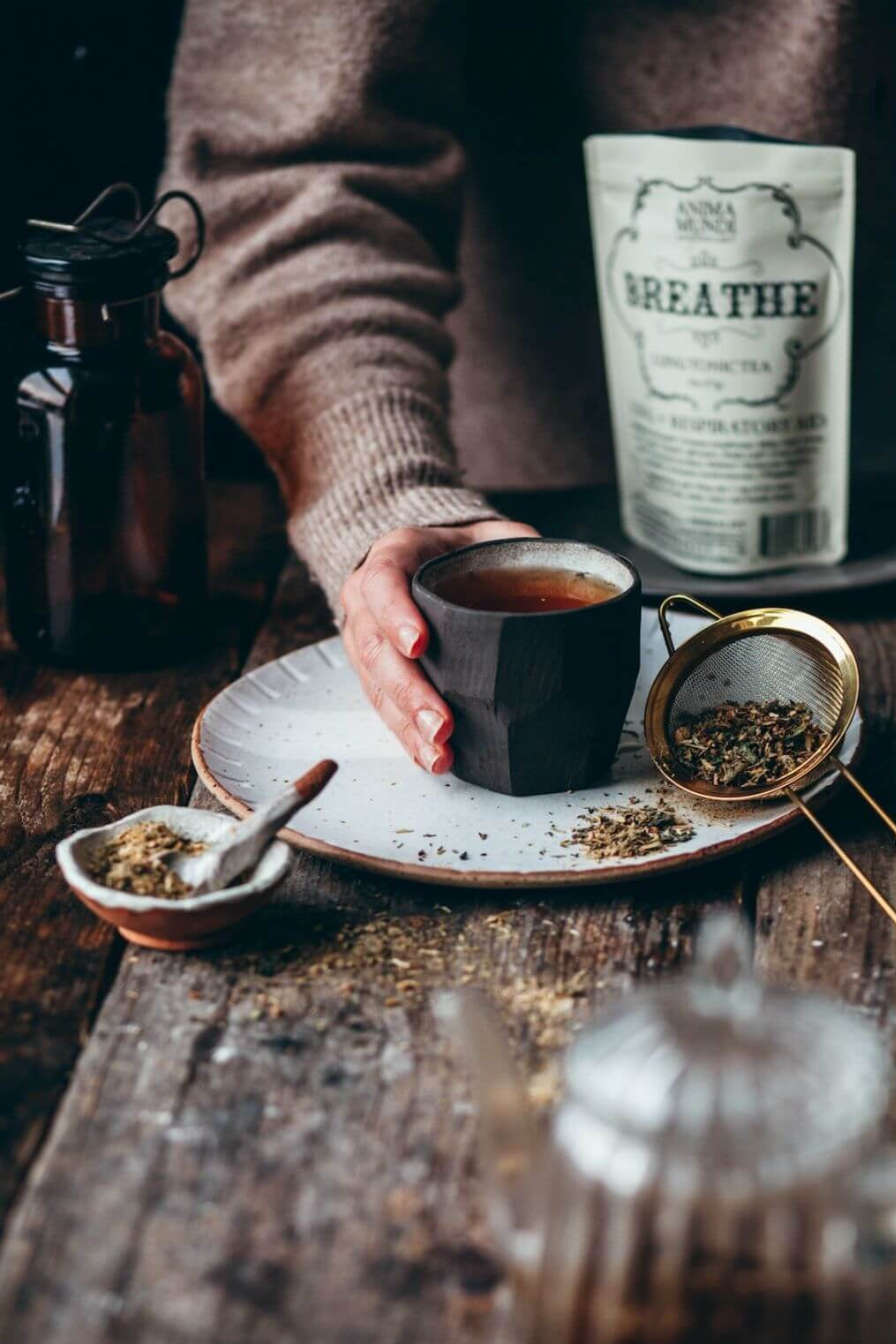 A person in a brown sweater holds a dark ceramic cup of tea on a speckled plate, with loose tea, a gold metal strainer, and a bag labeled 'BREATHE' on a rustic wooden table.