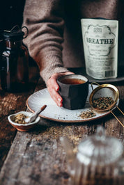 A person in a brown sweater holds a dark ceramic cup of tea on a speckled plate, with loose tea, a gold metal strainer, and a bag labeled 'BREATHE' on a rustic wooden table.