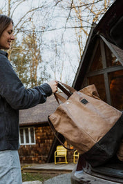 Woman loading waxed canvas tote bag into vehicle trunk beside cabin