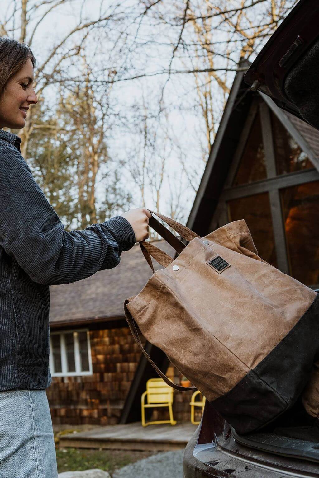 Woman loading waxed canvas tote bag into vehicle trunk beside cabin